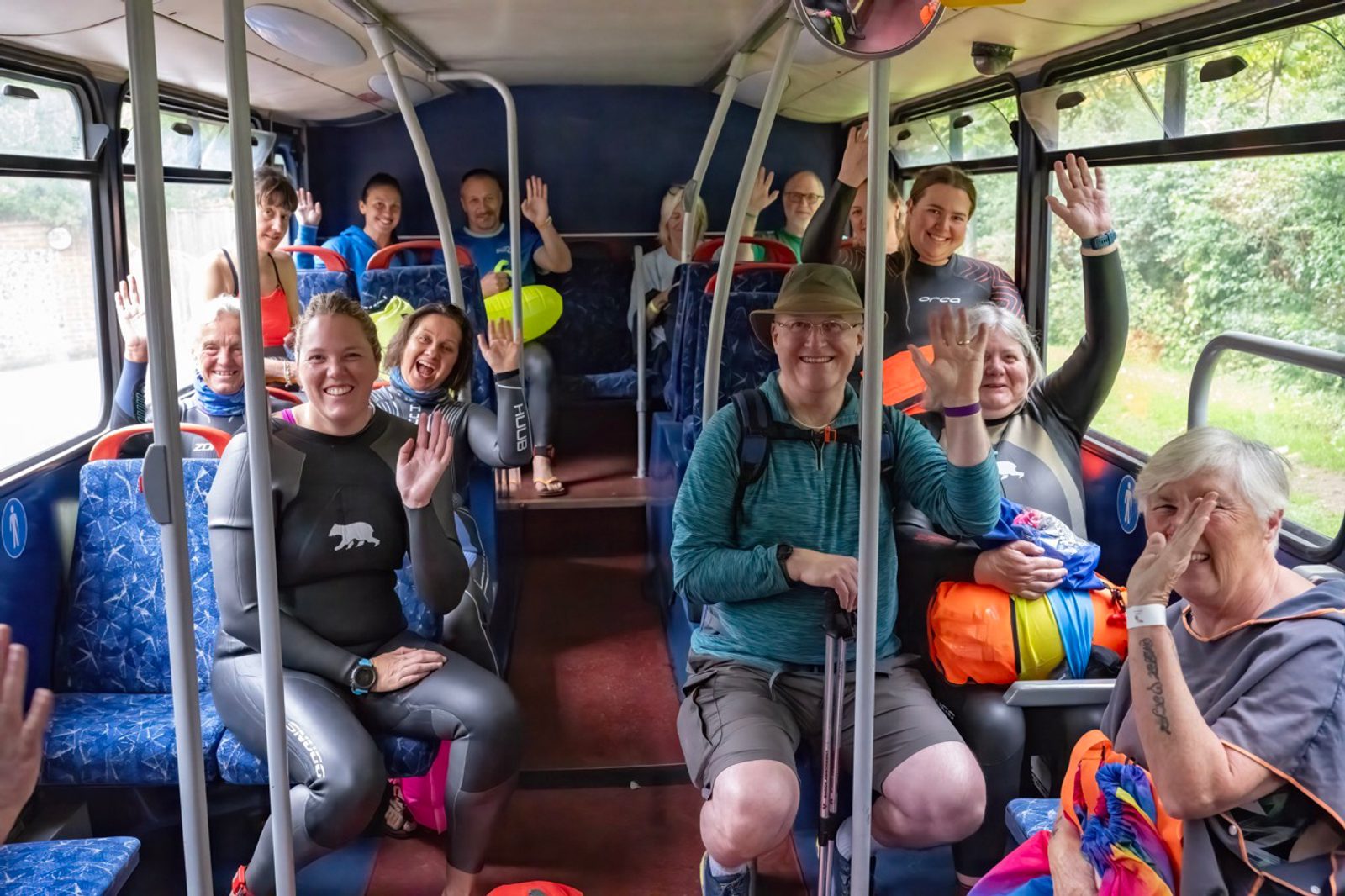 A group of smiling swimmers in wetsuits sit on a bus, waving towards the camera. They appear cheerful and ready for their activity. The bus interior has blue seats, and the outside greenery is visible through the windows.