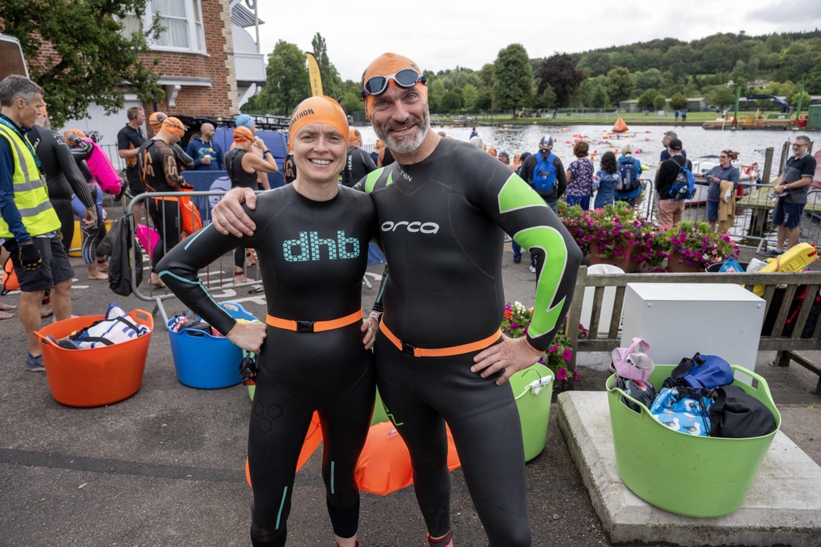 Two swimmers in wetsuits and orange swim caps pose for a photo in front of a busy outdoor swimming event. The woman on the left wears a black dhb wetsuit, and the man on the right wears a black Orca wetsuit. Others prepare for the event in the background.
