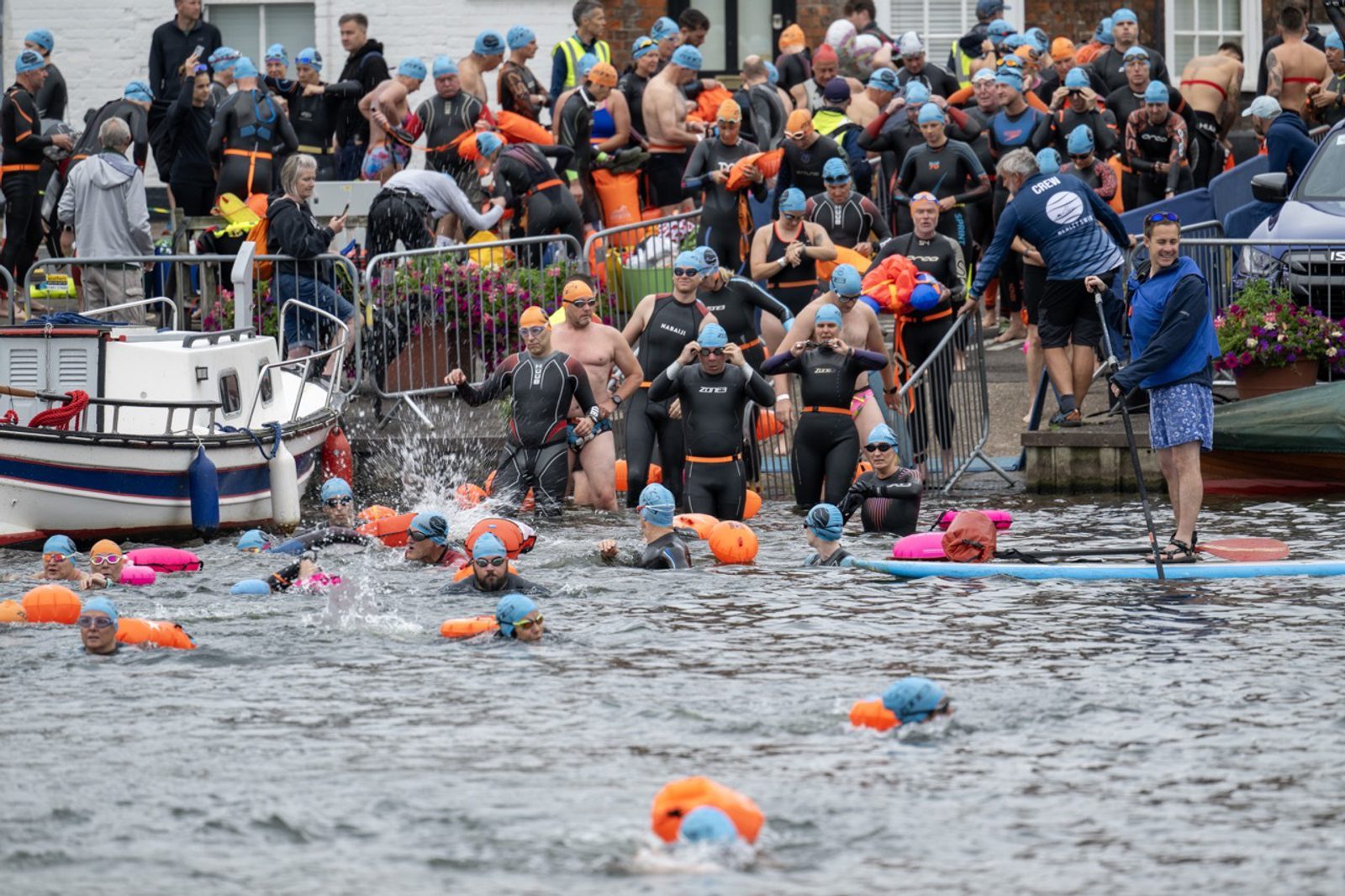 A large group of swimmers in wetsuits and colorful swim caps gather at the edge of a body of water, preparing for a swim event. Spectators and organizers are seen on the shore, some holding equipment, while a person on a paddleboard appears to give instructions.