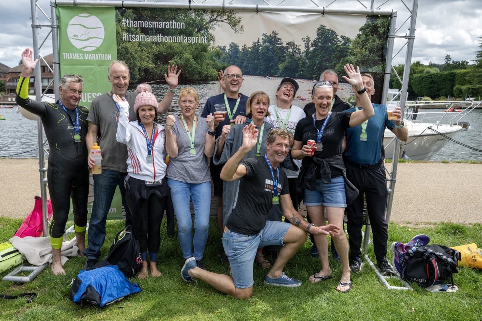 A group of people pose happily for a photo in front of a "Thames Marathon" banner, celebrating their participation. Some wear medals around their necks and athletic attire. Behind them is a scenic riverside with boats on the water and trees in the background.