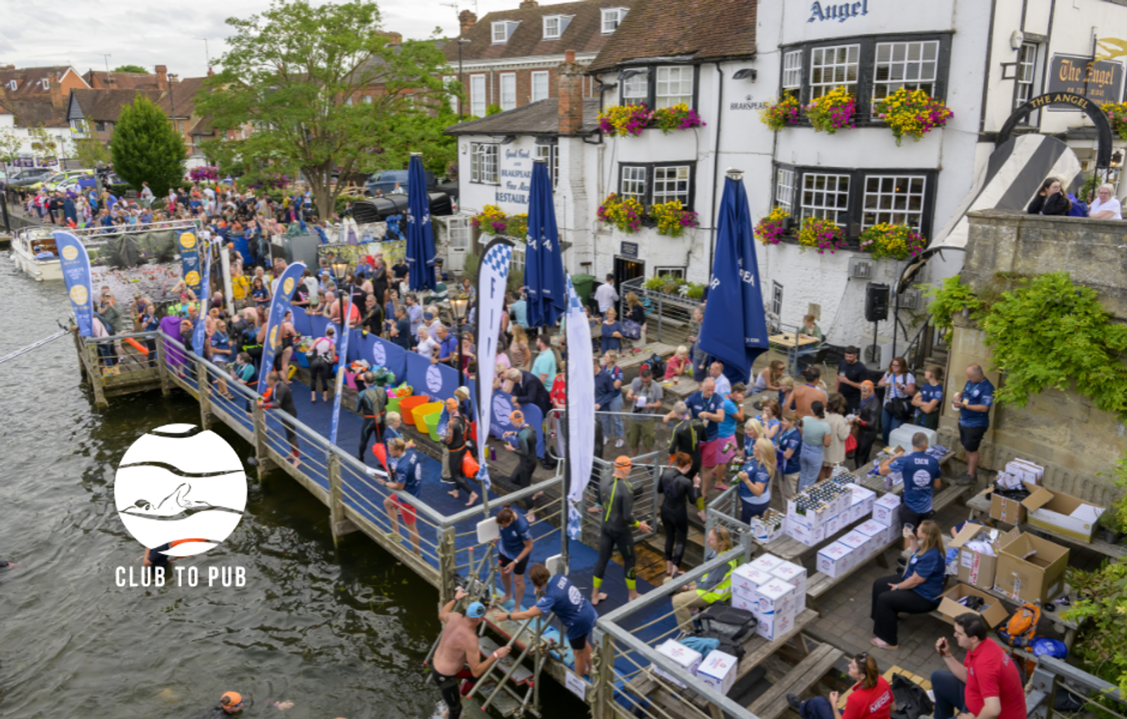 A bustling outdoor event by the riverside near a historic-looking pub. People are gathered around, many wearing blue shirts. Some participants are in swimming gear, while others are standing on the dock or mingling. Event banners and tables with supplies are visible.