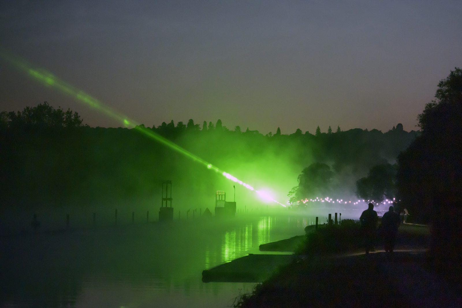 Silhouetted figures walk along a path beside a calm river at dusk. A vibrant green laser light pierces through the mist, creating an ethereal glow across the water. Trees and vegetation are faintly visible in the background, under a twilight sky.