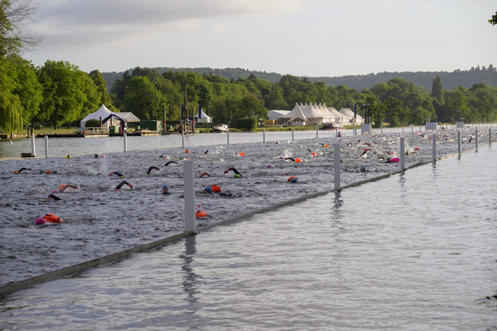 Swimmers in colorful caps participate in a competitive open-water race in a river, enclosed by white buoys, with green, tree-lined banks and tents in the background under a partly cloudy sky.