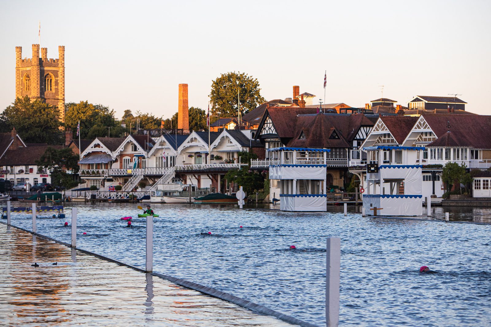 A scenic view of a river with swimmers participating in an event. The riverside is lined with charming, historic buildings. In the background, a church tower rises above the trees, under a clear sky.