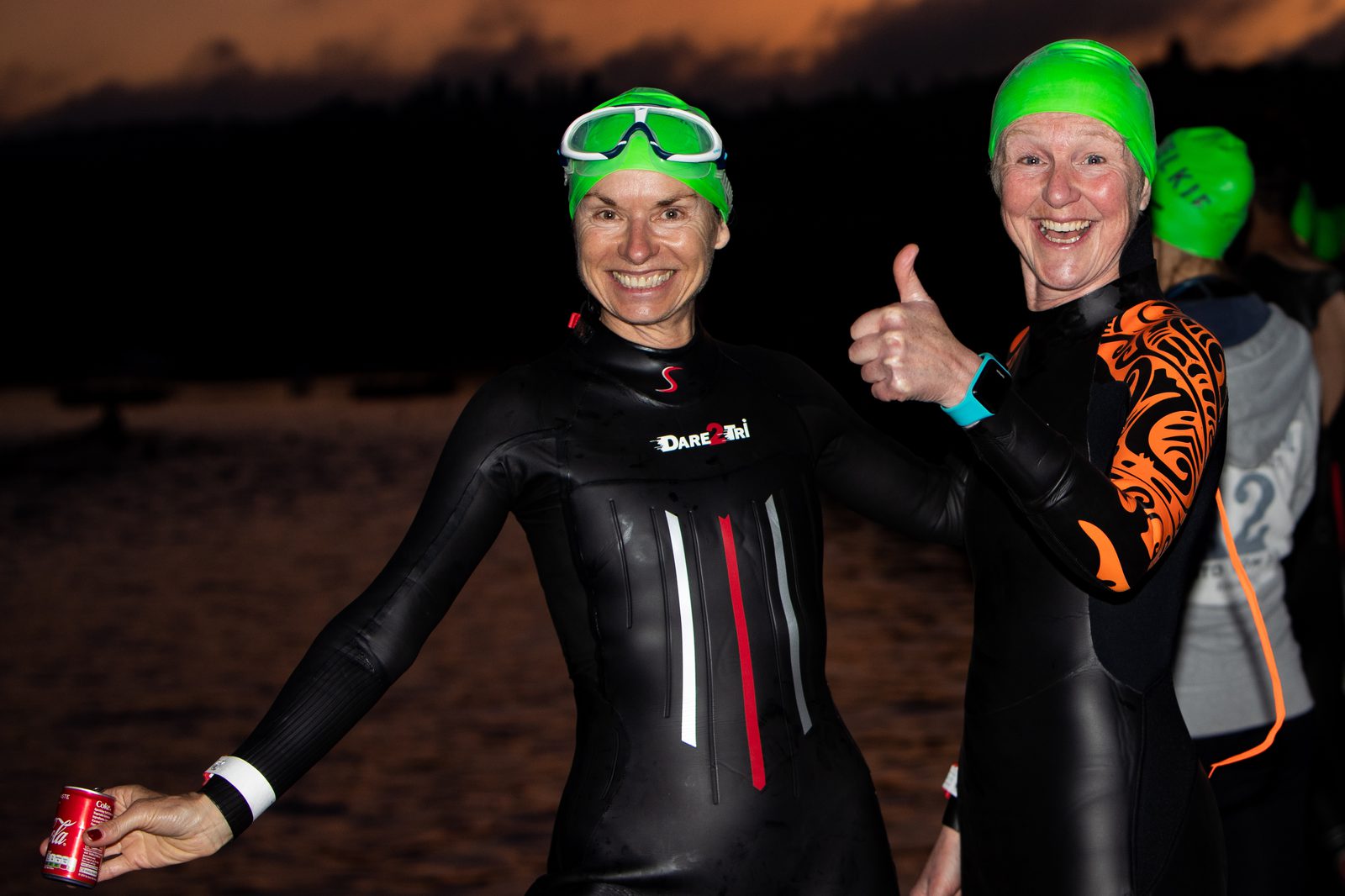 Two women in wetsuits, neon green swim caps, and goggles smile for the camera against a dark backdrop, possibly at dawn or dusk. One woman holds a small red can and the other gives a thumbs-up, indicating excitement and readiness for a swimming event.