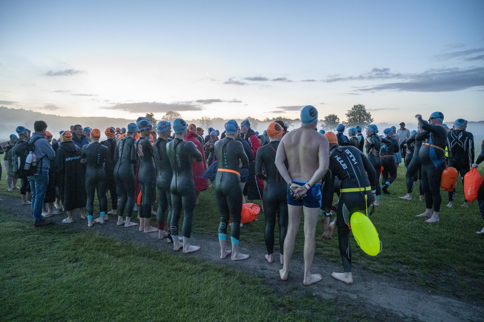 A large group of triathletes in wetsuits and swim caps, with safety buoys attached, gather on a grassy area near the water's edge early in the morning. The sky is lightening with the approaching dawn, and a few spectators stand nearby.