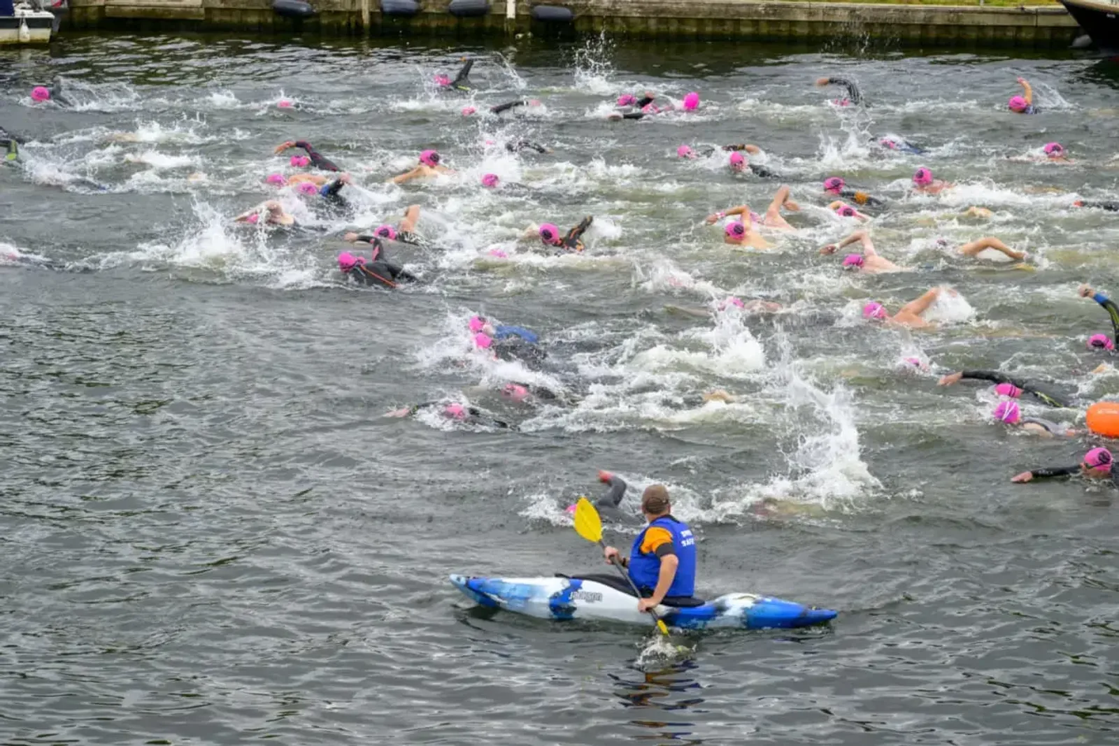 A group of people wearing pink swim caps participate in an open water swim race. The water is choppy from their movements as they swim towards a distant bank. A person in a blue and yellow kayak observes and guides the swimmers.