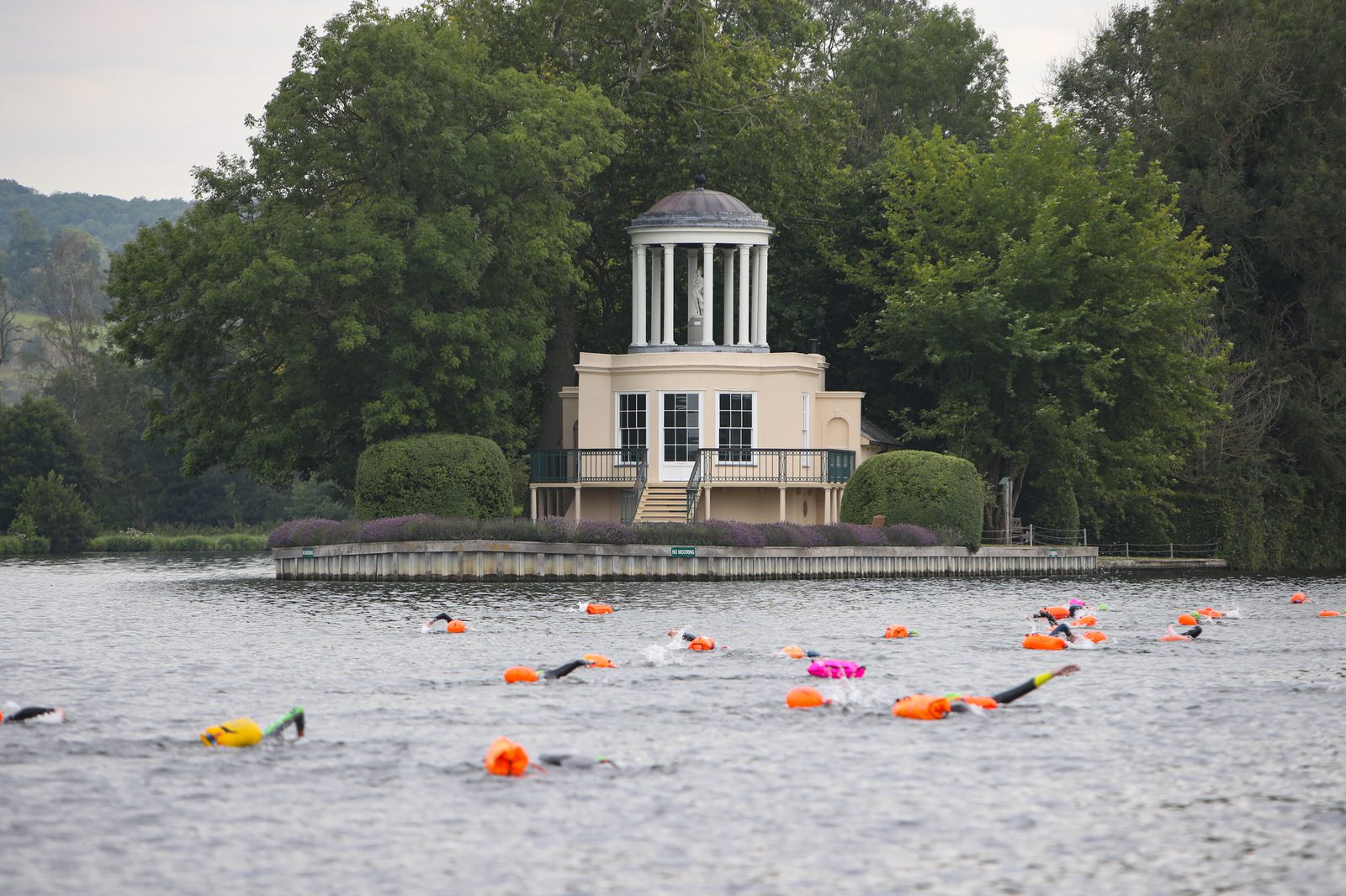 A riverside pavilion with a rotunda and large windows stands among lush green trees. In the river in front, numerous swimmers in brightly-colored swim caps and buoy markers participate in an open water swimming event.