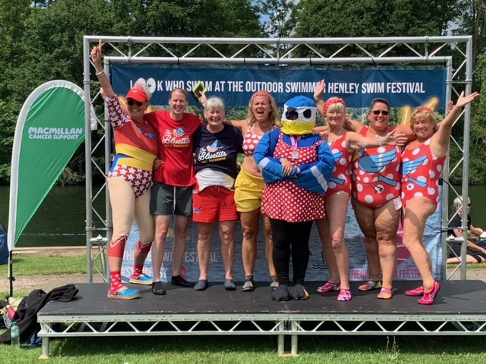 A group of eight people, some in colorful swimsuits and one in a duck costume, pose cheerfully with arms raised on a stage at an outdoor event. A green Macmillan Cancer Support banner is visible on the left, and greenery can be seen in the background.