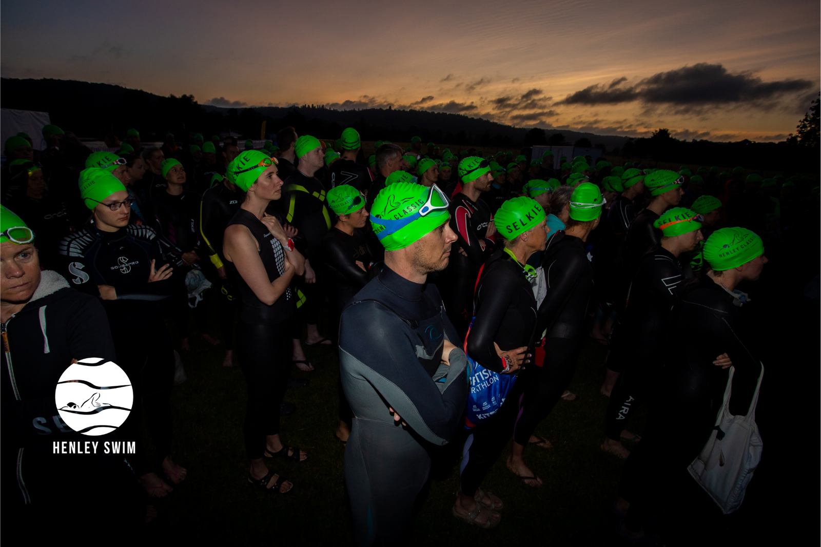 A large group of swimmers wearing black wetsuits and bright green swim caps stand together in a dimly lit outdoor area, some with arms crossed, as they prepare for a swim event at dawn. The sky is dark with the first light of the morning appearing on the horizon. A logo in the corner reads "Henley Swim.