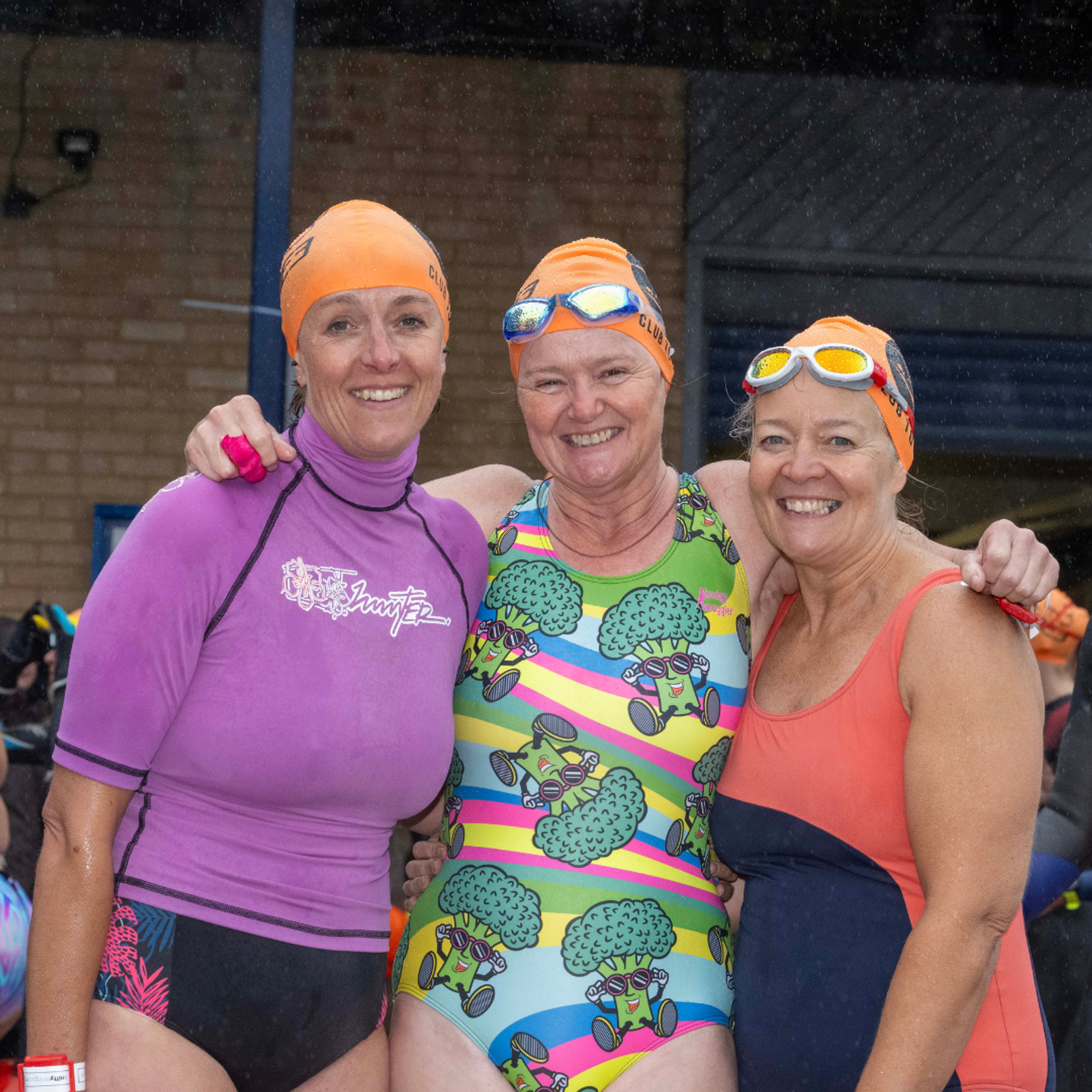 Three women wearing colorful swimsuits and orange swim caps with goggles smile at the camera, standing close together. The background shows a brick wall and some outdoor structures, suggesting they are at a swimming event.