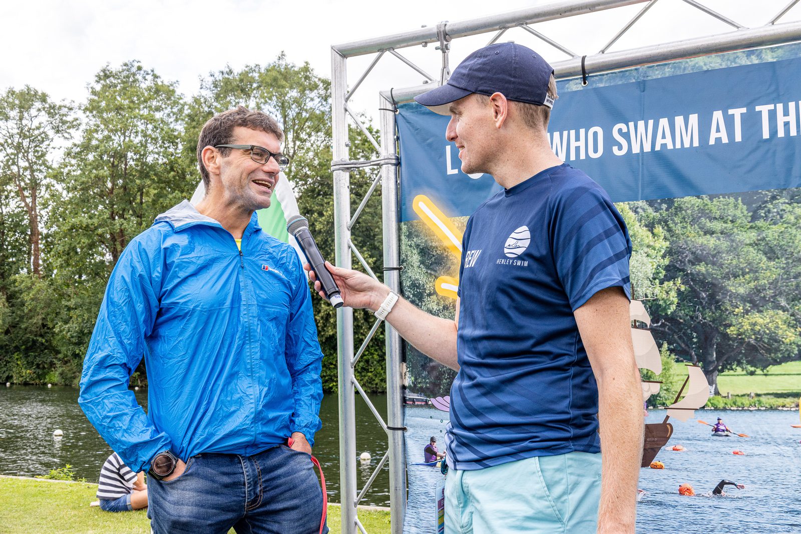 Two individuals are having a conversation outdoors near a lakeside with a "Who Swam at the" sign partially visible in the background. The person on the left, wearing glasses and a blue jacket, is speaking into a microphone held by the person on the right, who is donning a navy shirt and cap.