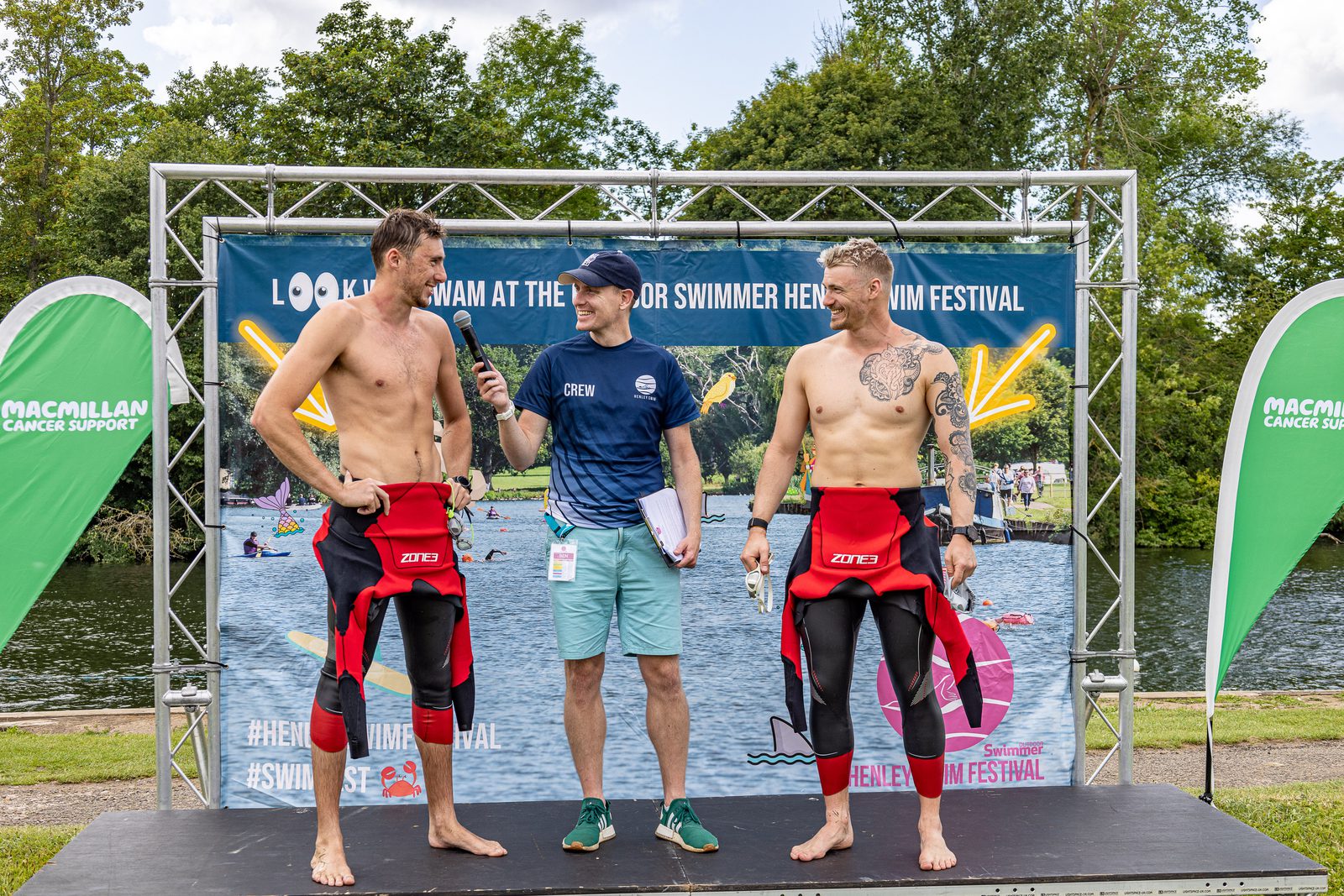 Three men are standing on a stage at an outdoor swim festival. The man in the center, wearing a shirt and cap with "crew" written on it, is interviewing two men in red and black wetsuits holding the top parts pulled down. A lake, trees, and banners are visible in the background.