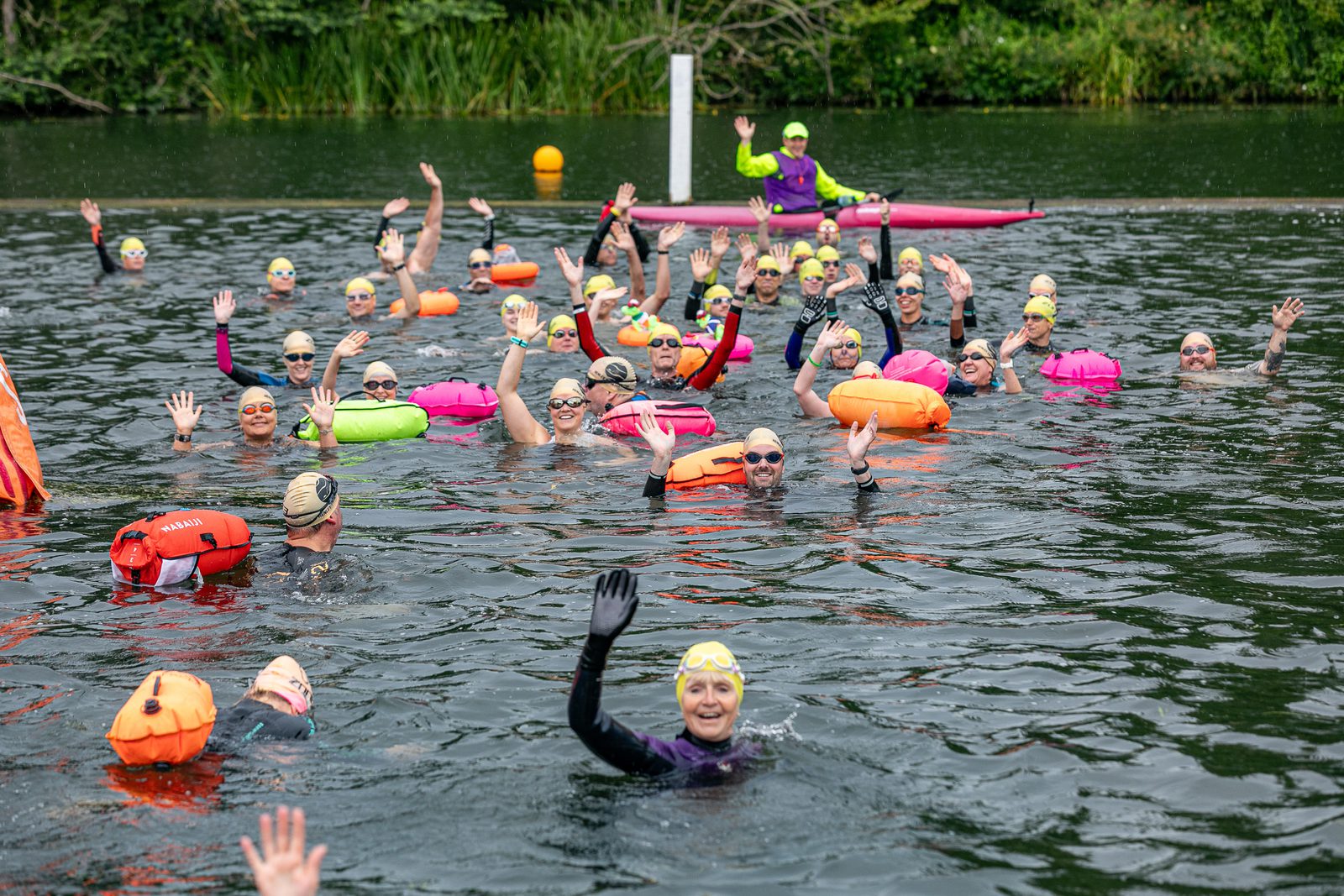 A group of people wearing swim caps and holding colorful buoyancy aids wave at the camera while standing in the water of a lake. They appear to be participating in an open-water swim event. A water safety official in a kayak is seen in the background.