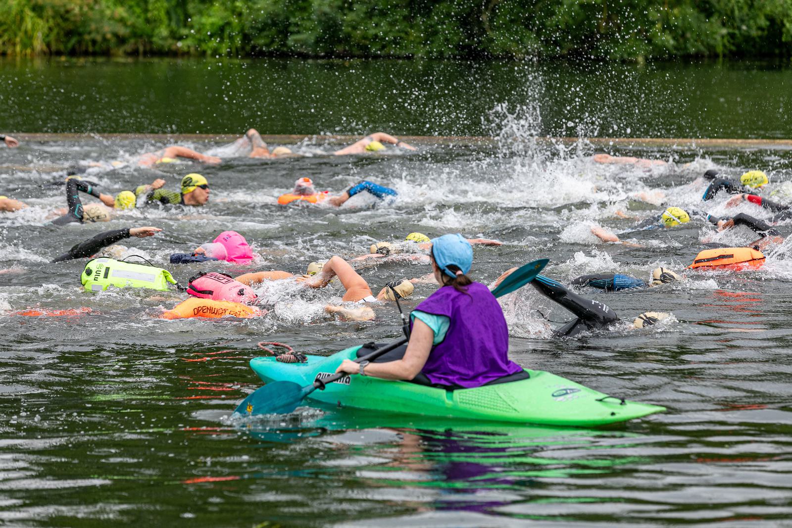 A group of swimmers wearing brightly colored swim caps and safety buoys participate in an open-water swimming race on a lake. A person in a green kayak, wearing a purple vest and blue cap, supervises the swimmers. The background is lush and green with trees.