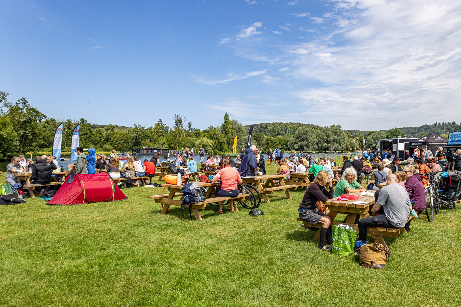 People are sitting at wooden picnic tables and on the grass, enjoying food and drinks at an outdoor event in a park. The sky is clear and blue, and there are various tents and banners in the background. The scene is lively, with diverse groups of people and various activities.