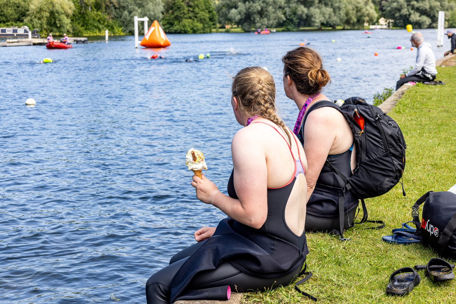 Two women in wetsuits sit on the grassy bank beside a river, watching activities on the water. One woman is eating an ice cream cone. Backpacks and other personal items are next to them. There are markers and kayaks on the river, and trees are visible in the background.
