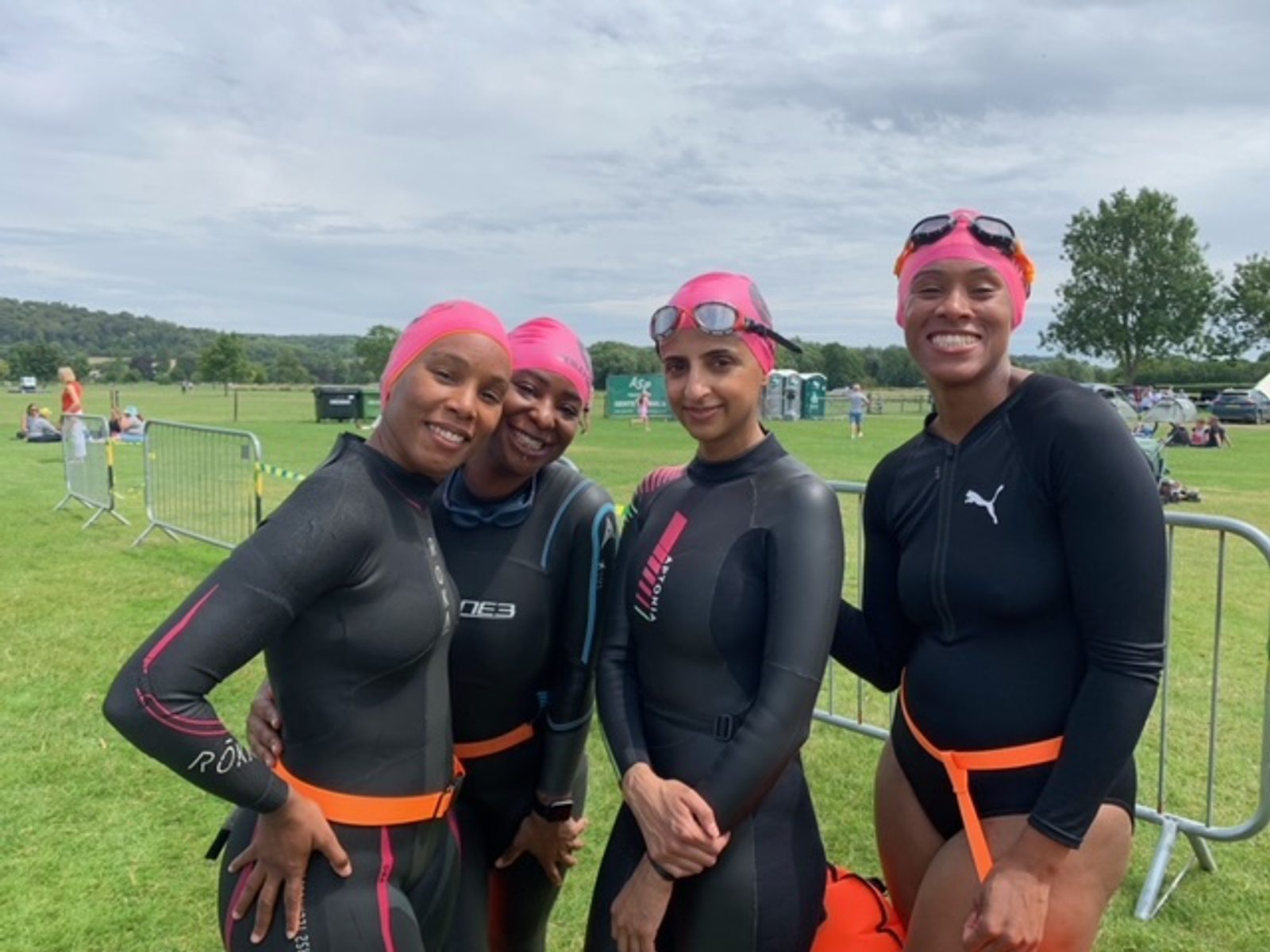Four women wearing wetsuits and pink swim caps are standing together and smiling at a grassy outdoor event. Safety barriers and other people are visible in the background. The sky is partly cloudy.