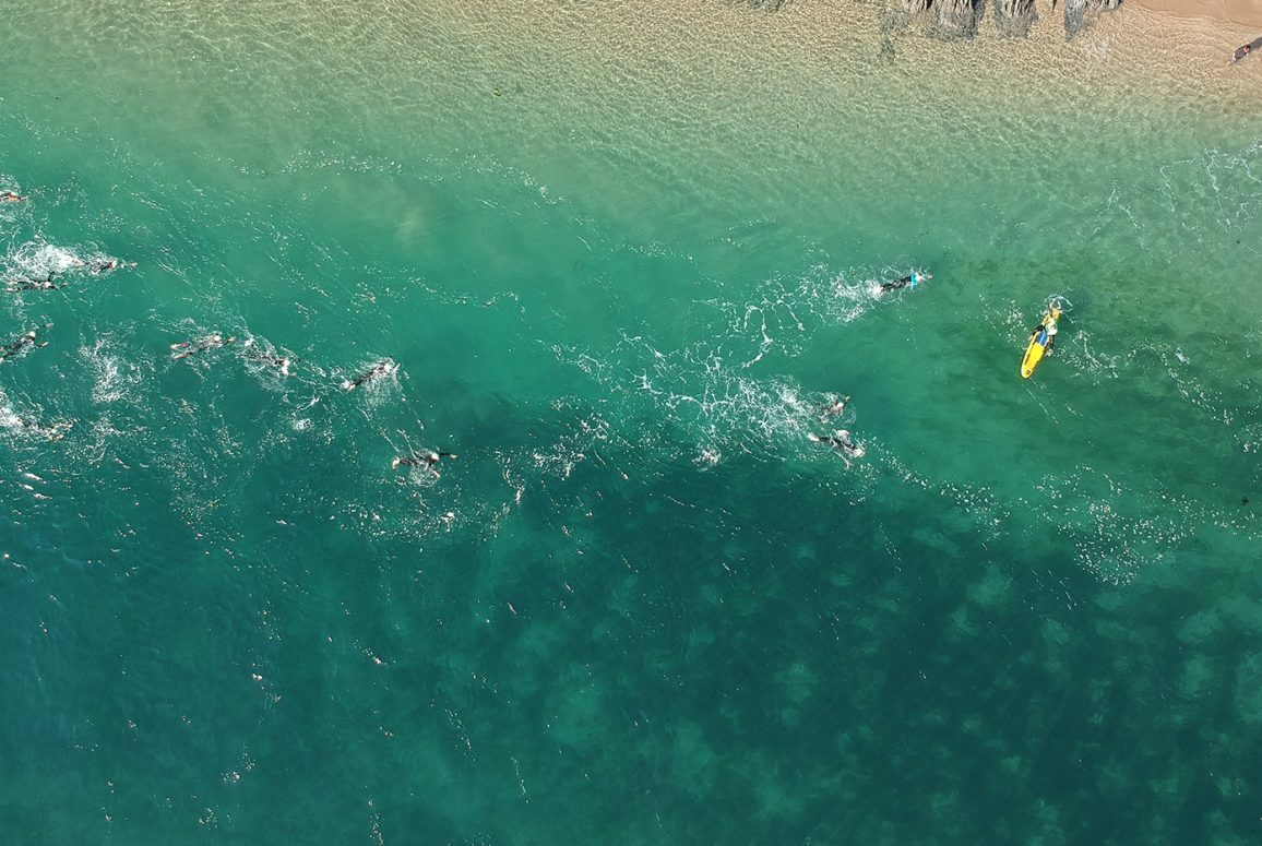 Aerial view of a group of people swimming in clear turquoise ocean water near a sandy beach. A person on a yellow float or surfboard is also visible among the swimmers. The scene captures an energetic and refreshing aquatic activity.