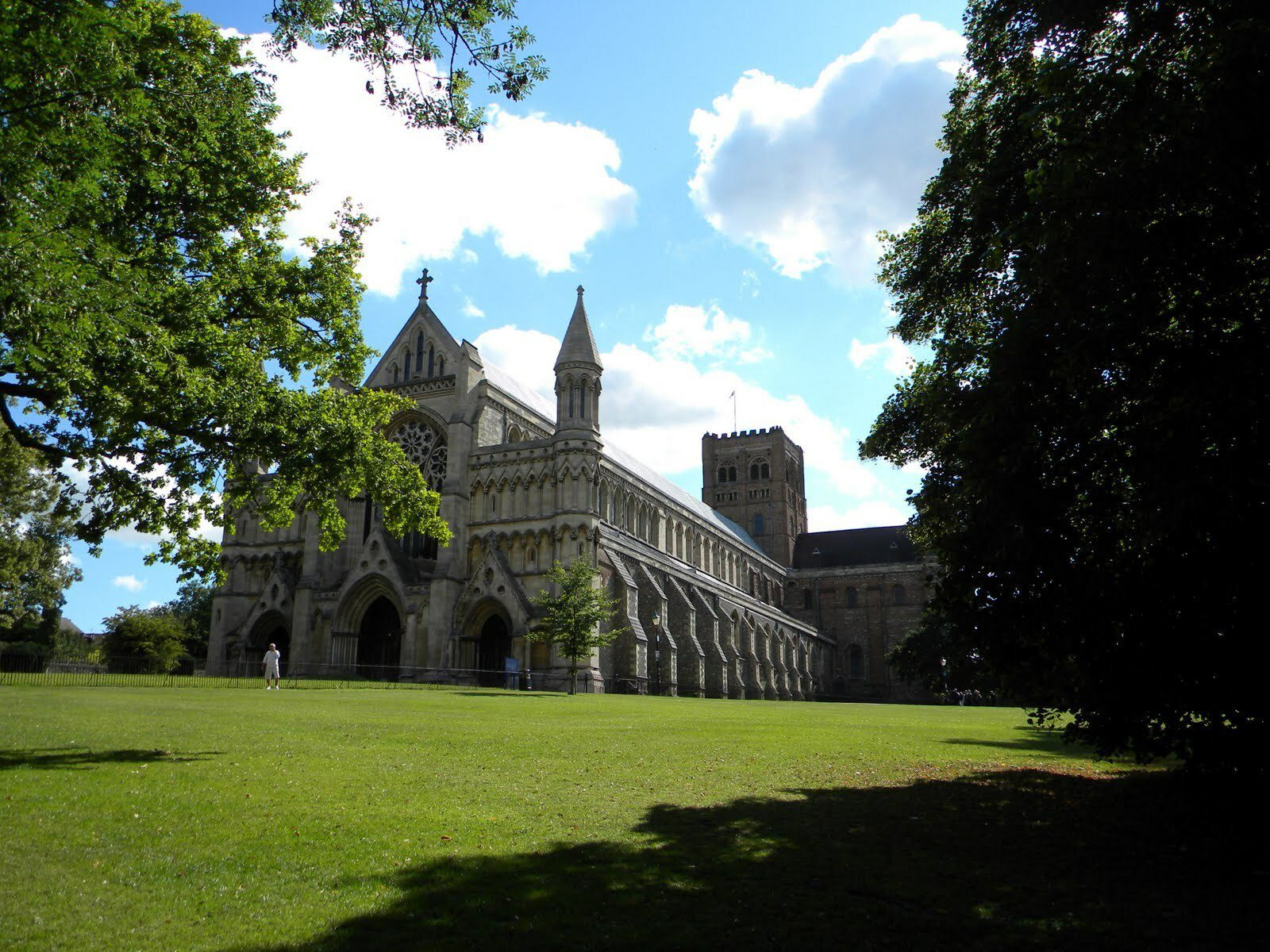 A historic stone cathedral with arched windows and twin spires is set against a backdrop of a bright blue sky with scattered clouds. Lush green grass and tall trees surround the cathedral, casting shadows on the lawn. A person walks near the building.