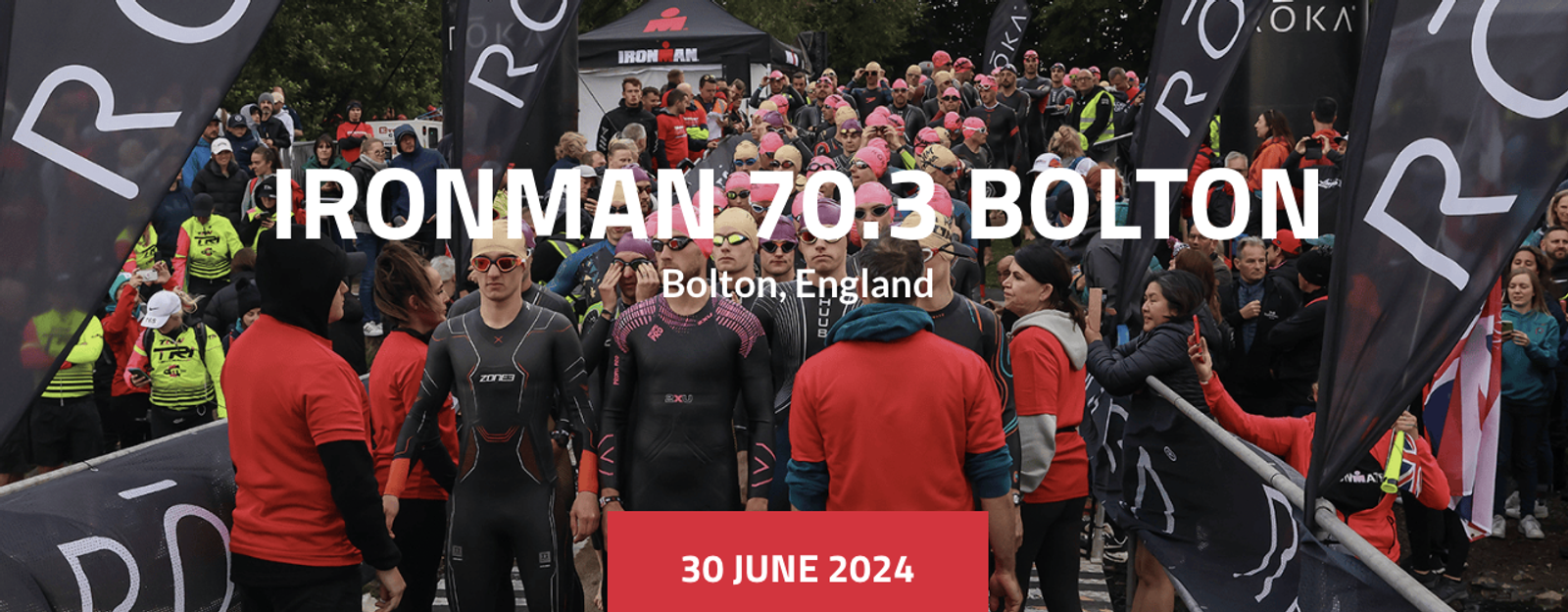 A large group of participants wearing wetsuits and swim caps stand at the starting area of the IRONMAN 70.3 Bolton triathlon in Bolton, England. Spectators and event staff are also gathered around. The date, 30 June 2024, is displayed at the bottom.