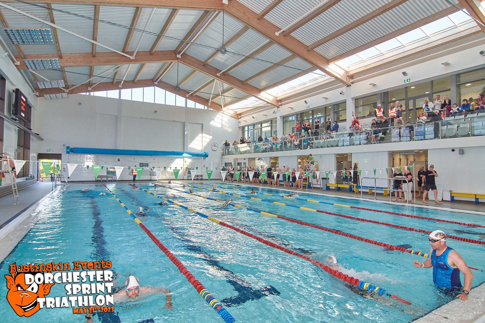 An indoor swimming pool with several swimmers participating in a race. Spectators observe from a balcony. Event banners and the logo for "Bustinskin Events Dorchester Sprint Triathlon May 1st, 2022" are visible in the bottom left corner.