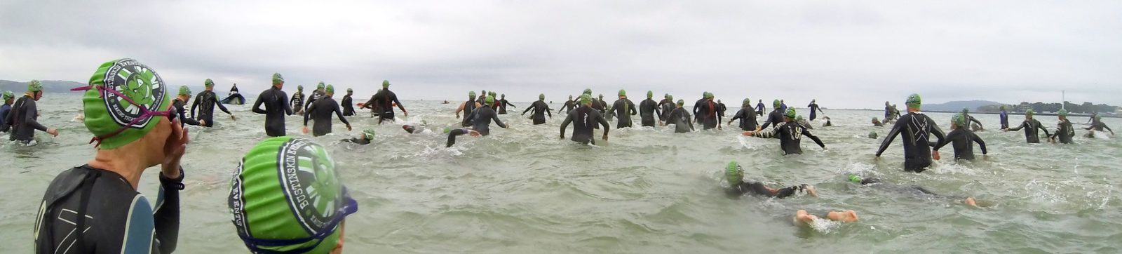 A large group of swimmers wearing black wetsuits and green swim caps are in the water, possibly participating in a triathlon or open water swim event. The scene is overcast with a distant view of the shoreline and some trees.