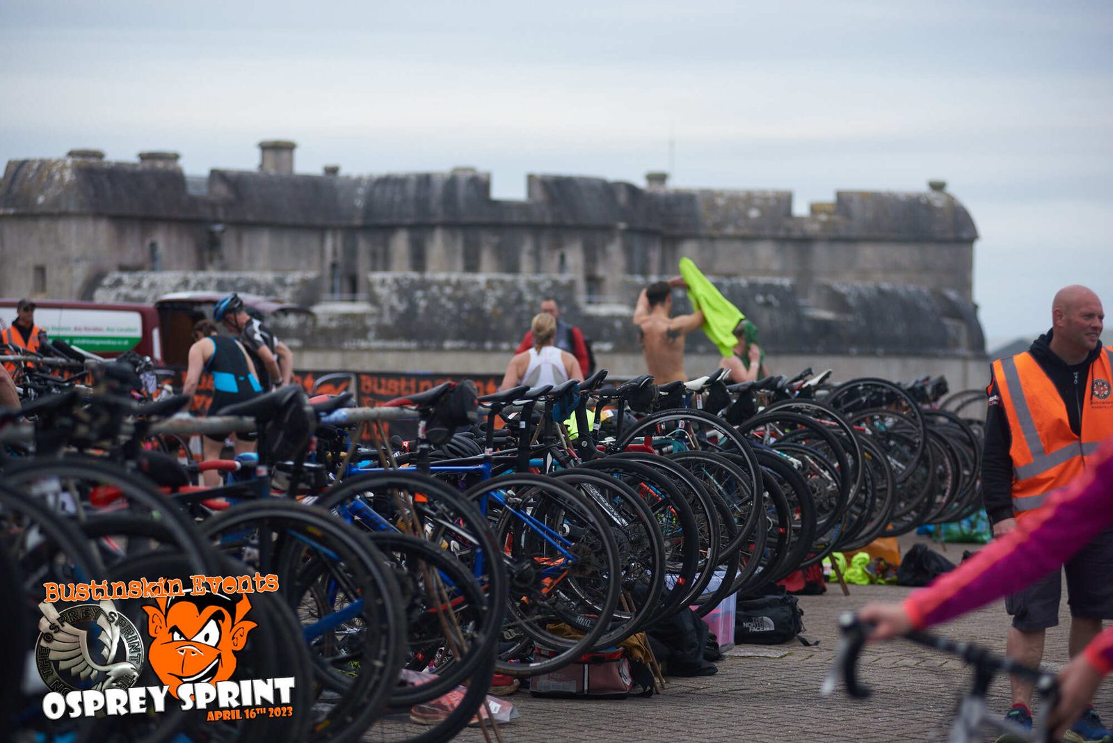 A group of bicycles is parked in a designated transition area during a sporting event. Participants and volunteers, some wearing orange vests, are visible. A large stone fort is in the background. The event's logo and details are displayed in the bottom left corner.
