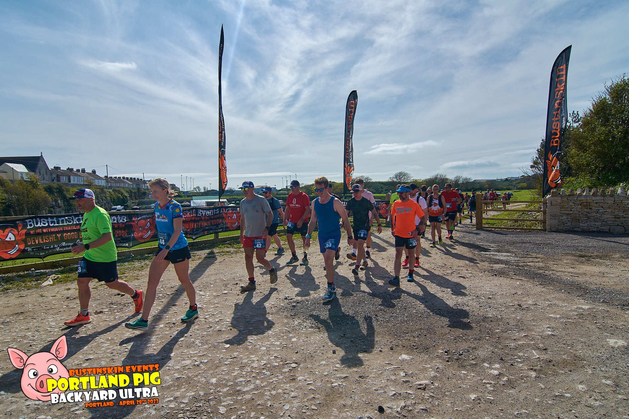 A group of runners at the starting line of the Portland Pig Backyard Ultra race, with colorful event flags and banners around them. The sky is clear, and the race appears to be taking place in a scenic, rural setting. The event logo is at the bottom left corner.