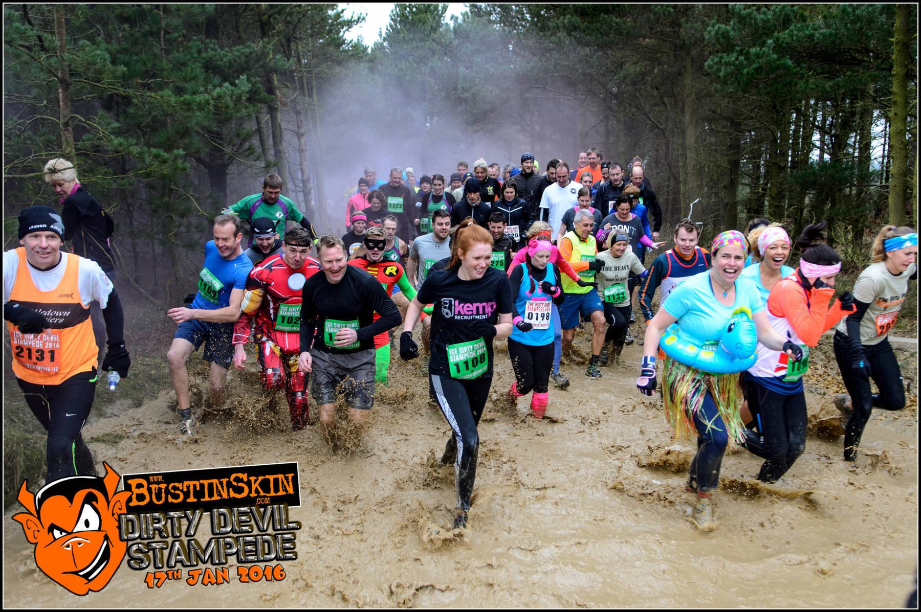 Participants in colorful outfits run through a muddy obstacle course in a forested area during the BustinSkin Dirty Devil Stampede on January 17, 2016. Some wear superhero costumes, tutus, and inflatable armbands, smiling and laughing as they race.
