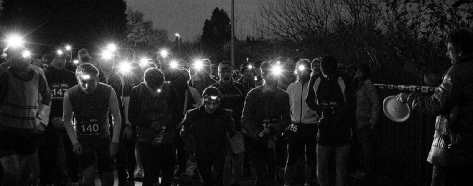 A group of runners, each wearing a headlamp, prepares for a night race. They are standing closely together in the dark, with trees and a few buildings faintly visible in the background. A person with a clipboard is on the right.