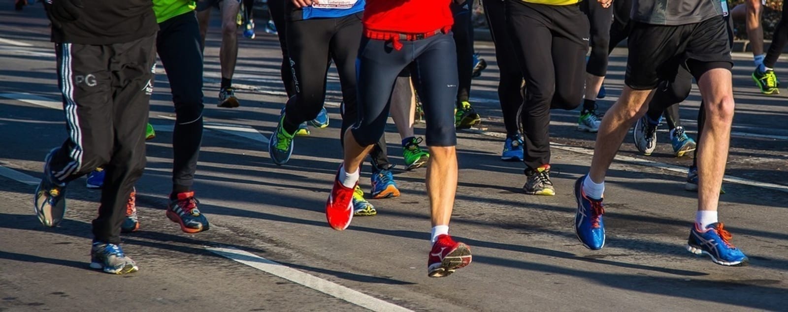 Close-up view of the legs of a group of runners in athletic clothing and various colorful running shoes participating in a road race on a paved street. The runners' torsos are mostly out of the frame, emphasizing the motion of their legs and feet.