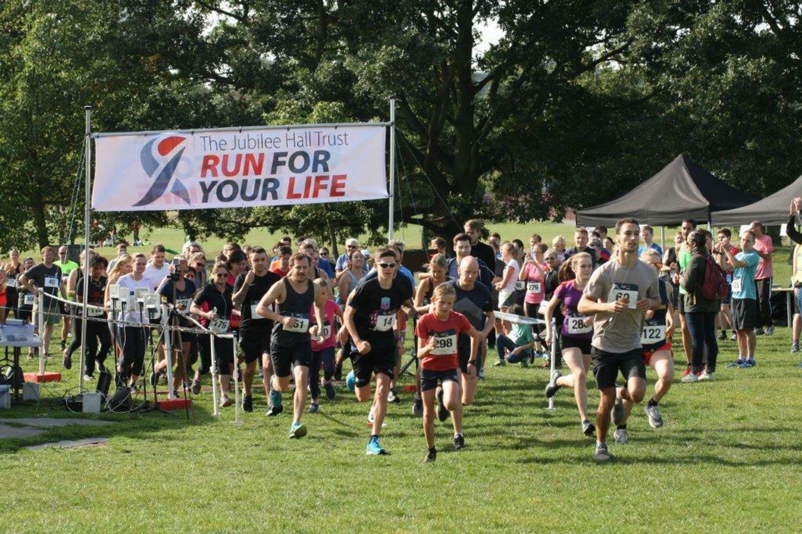A large group of runners of various ages are starting a race outside on a grassy field. They are running under a banner that reads "The Jubilee Hall Trust Run For Your Life." Spectators and tents are visible in the background under clear skies.