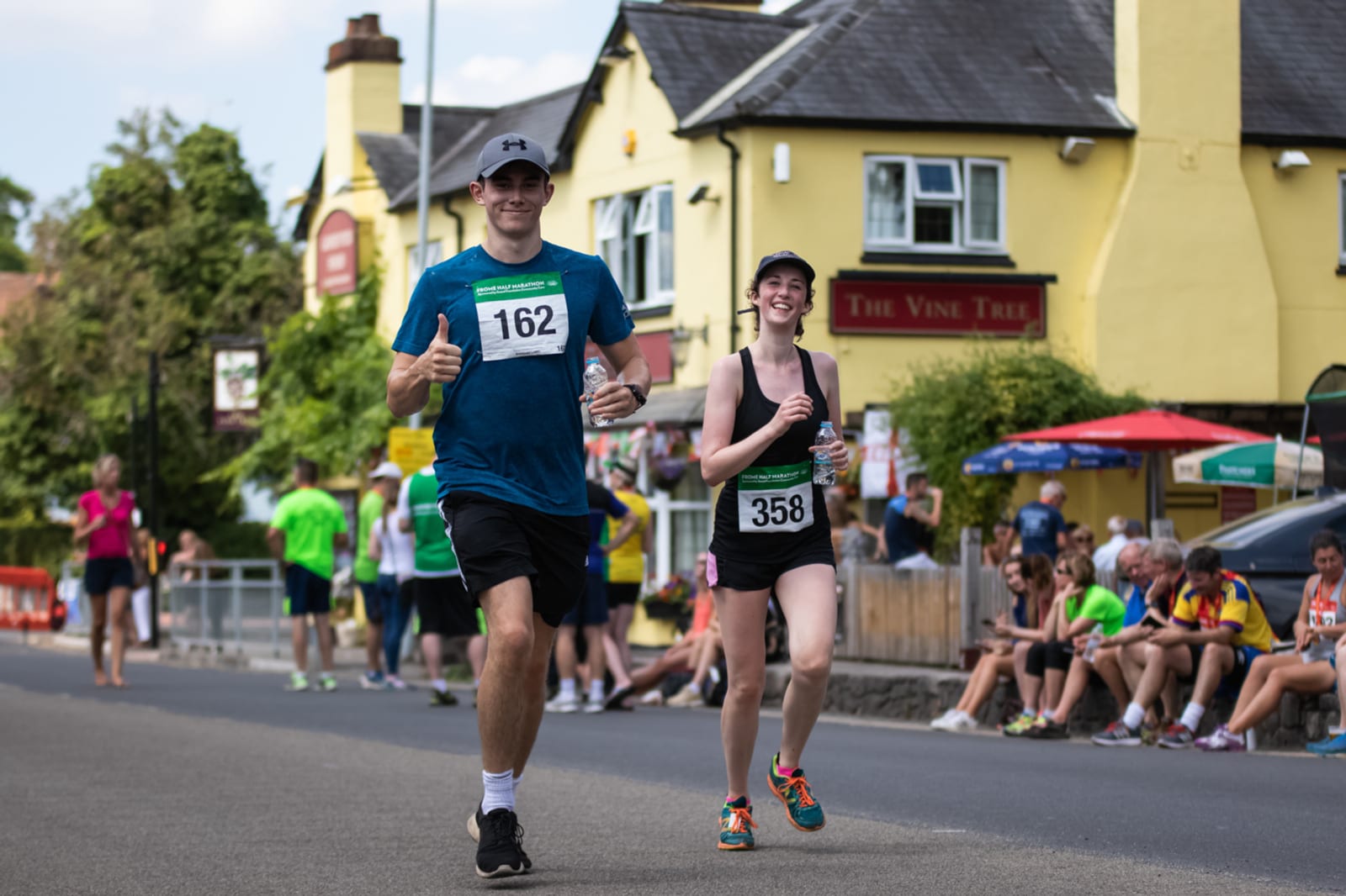 Two runners, one wearing a blue shirt and bib number 162, and the other wearing a black tank top and bib number 358, are smiling and giving thumbs up during a race. They are on a street with spectators sitting and standing in front of a building with a sign that reads "The Vine Tree.