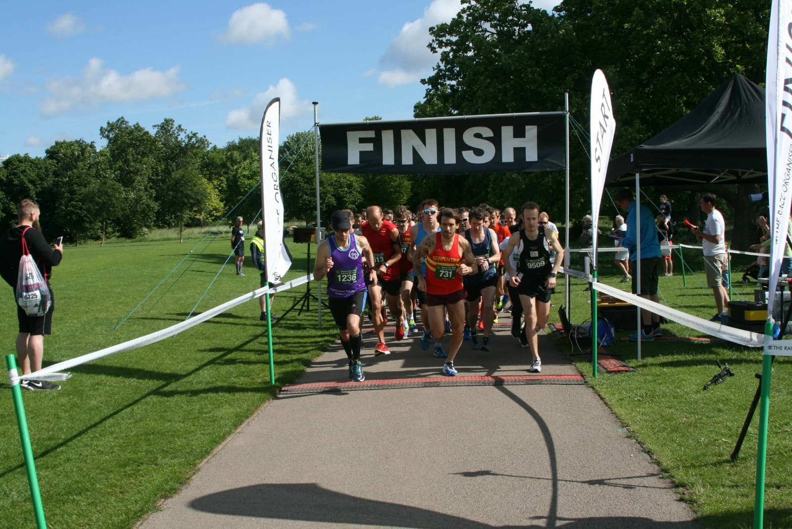 A group of runners crossing the finish line of a race on a paved path in a park. Some runners wear numbered bibs and various athletic gear. Blue sky and green trees are in the background. Spectators can be seen to the left and right of the finish line.