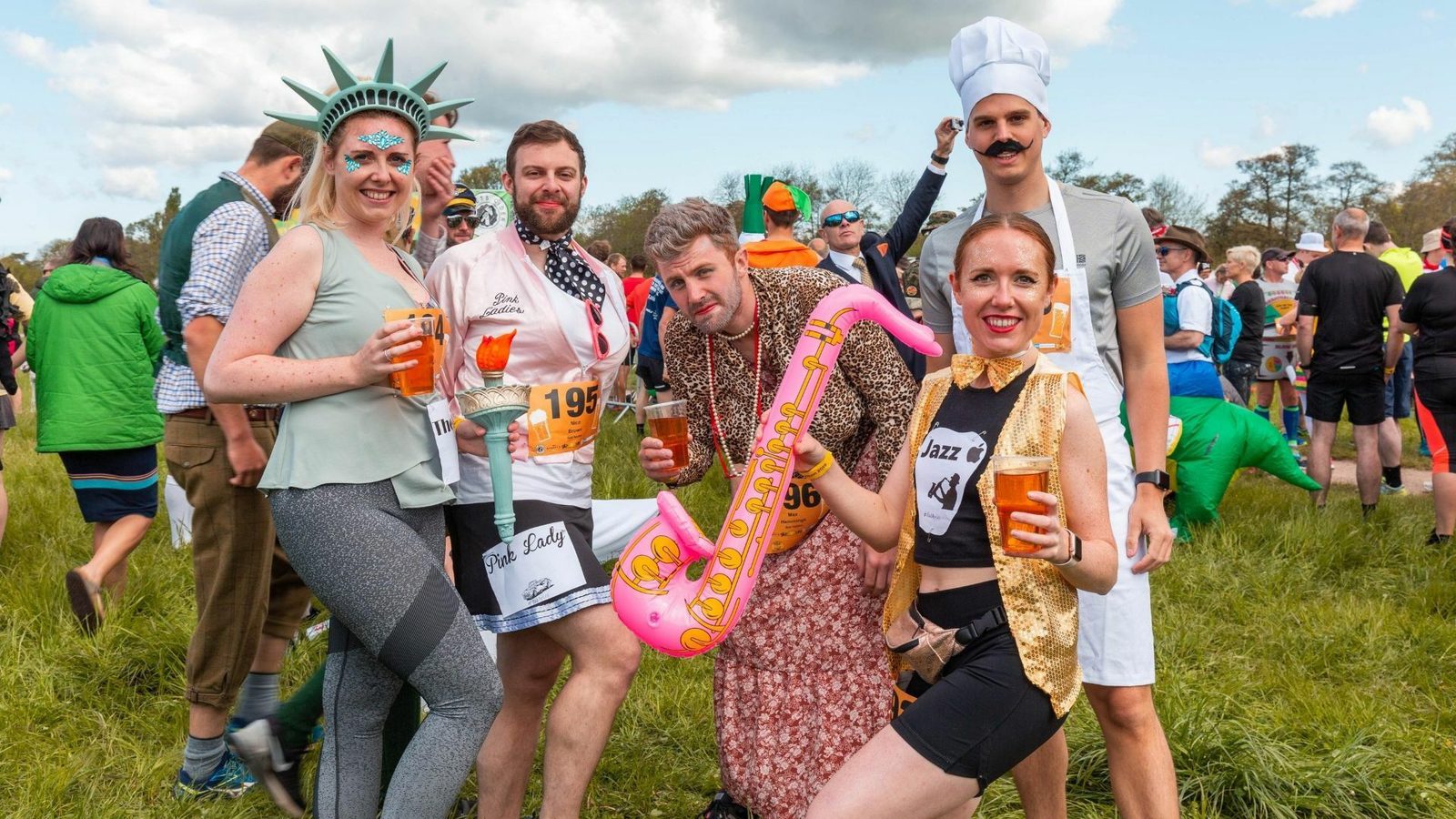 A group of people in costume poses outdoors. One person is dressed as the Statue of Liberty, another as a chef, one wears a moustache and chef's hat, and others have various whimsical outfits like jazzy attire and holding an inflatable saxophone. They all hold drinks.
