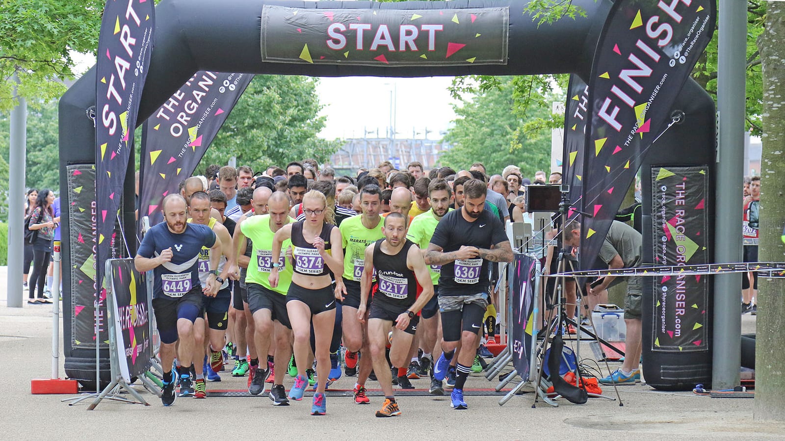 A group of runners, dressed in sportswear, begin a race at the starting line under a large black "START" arch with matching "FINISH" banners on each side. The scene is lively, with diverse participants focused and in motion. Trees and spectators are visible in the background.
