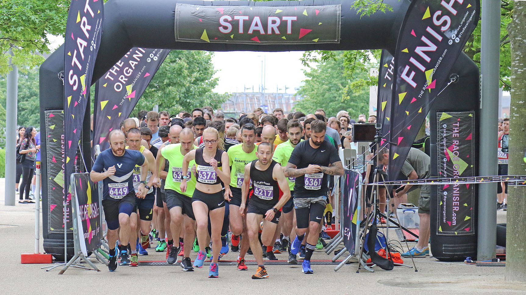 A group of runners, dressed in sportswear, begin a race at the starting line under a large black "START" arch with matching "FINISH" banners on each side. The scene is lively, with diverse participants focused and in motion. Trees and spectators are visible in the background.