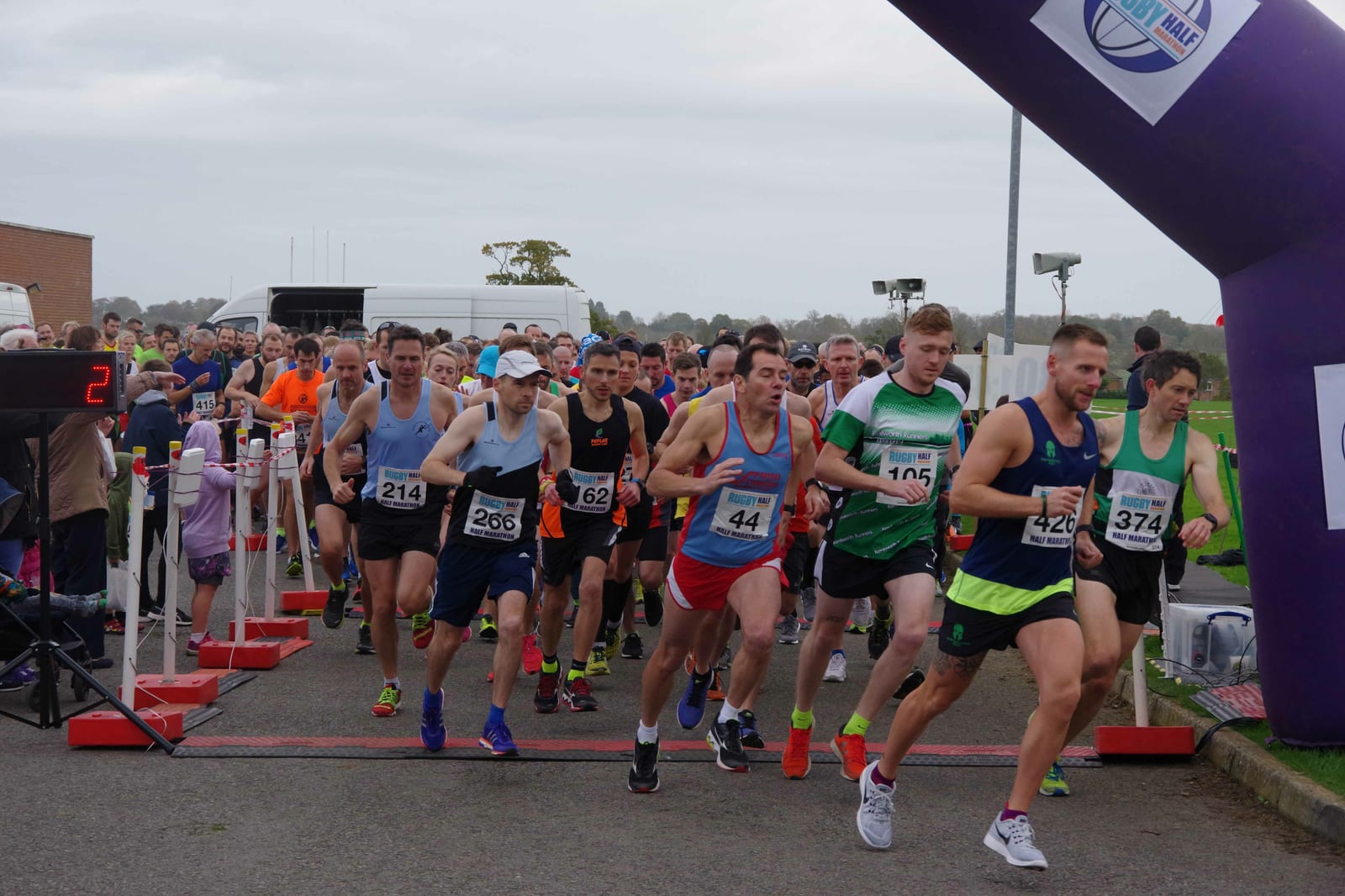 A group of runners starting a race under an inflatable archway. The participants are wearing various athletic outfits with race bibs pinned to their shirts. Spectators and volunteers are visible in the background. The sky is overcast.
