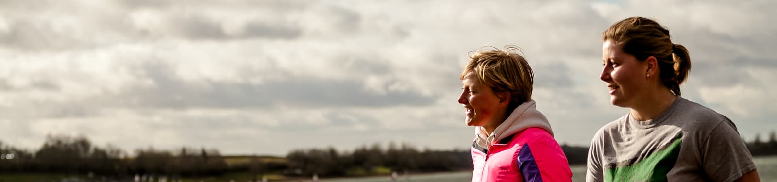 Two women are outdoors, standing side by side and smiling. They appear to be engaged in conversation. One is wearing a bright pink jacket, and the other is wearing a gray shirt. The background shows a cloudy sky and some distant trees.