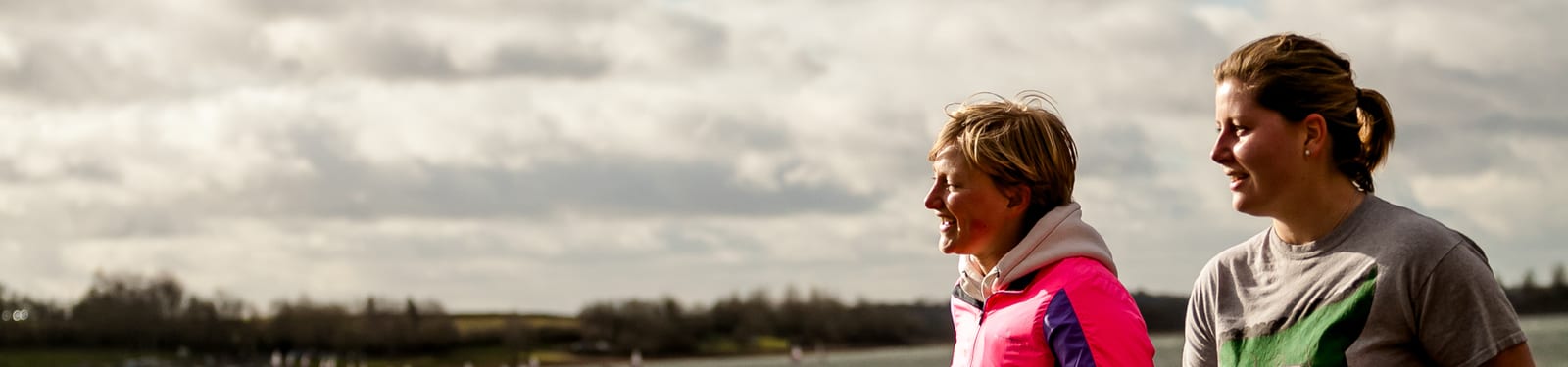Two people stand outdoors under a cloudy sky. The person on the left wears a bright pink jacket, while the person on the right wears a gray shirt. Both are smiling and appear to be engaged in conversation. A landscape with trees and fields is visible in the background.