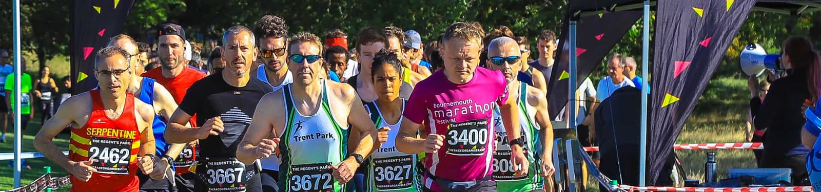 Runners at a starting point participate in a marathon. They are tightly packed as they begin the race, some in brightly colored athletic gear, with bib numbers visible. Trees and event tents are in the background under a clear blue sky.