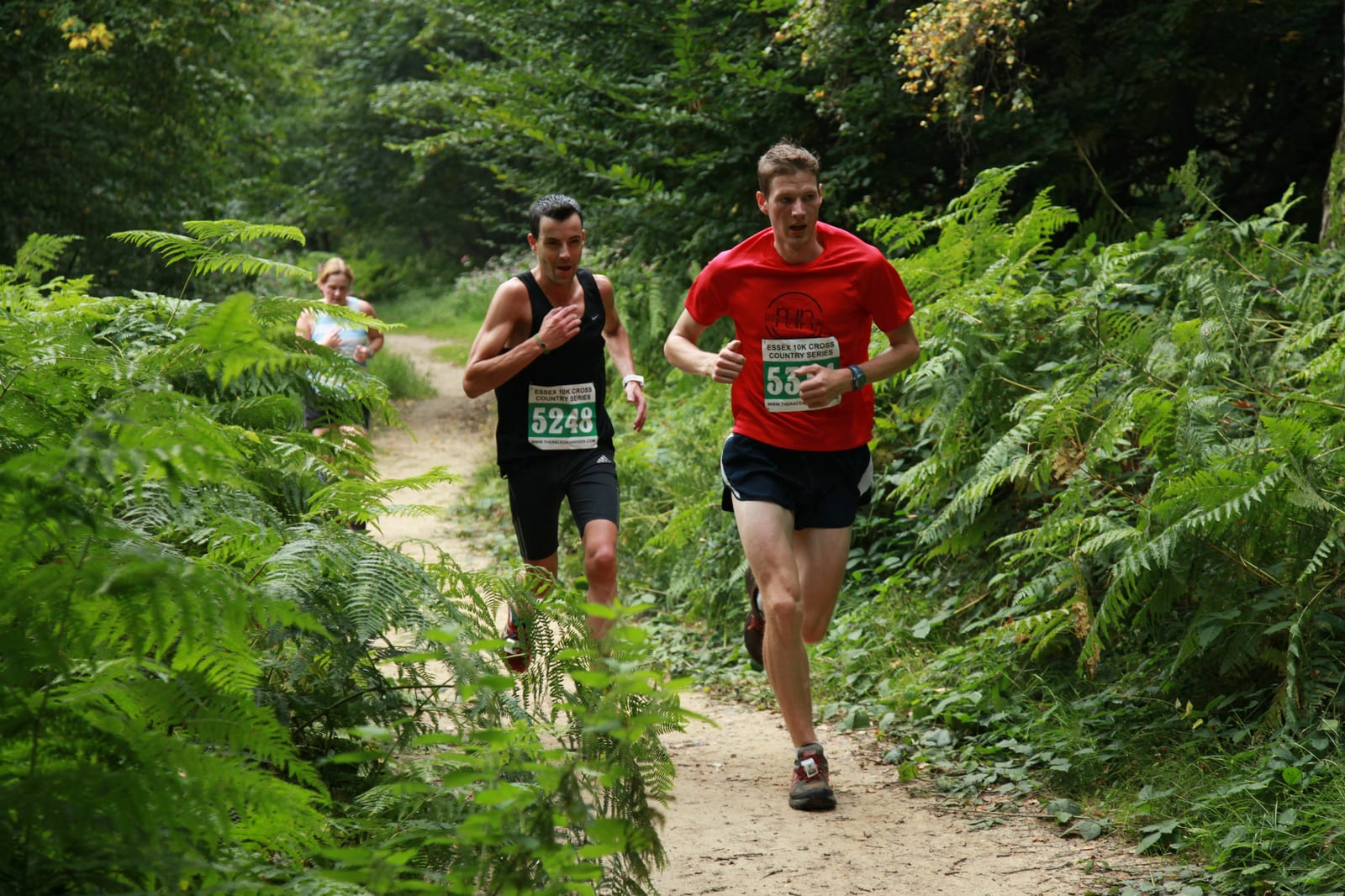 Three people running in a forest trail race surrounded by lush green ferns and trees. The runner in the red shirt leads the group, followed closely by a runner in a black tank top, with a third runner visible further behind.