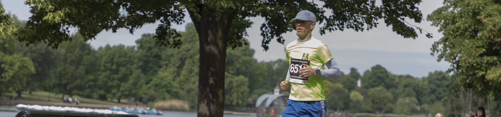 A person wearing a yellow shirt, blue shorts, white arm sleeves, and a gray cap is running in a park. The park features a large tree in the foreground and a pond with boats in the background. The person is wearing a bib labeled "253.