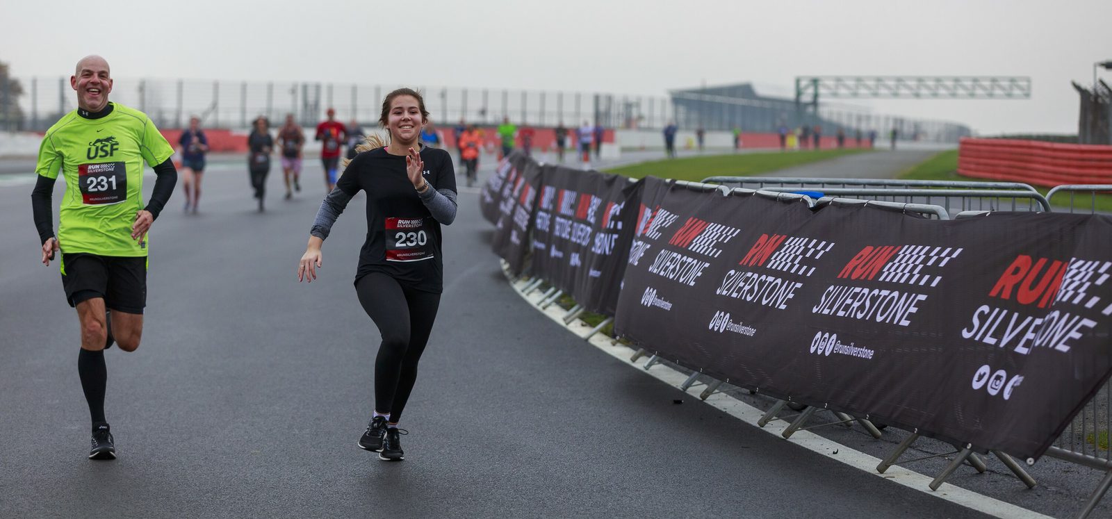 A group of runners on a racetrack, with two individuals in the foreground smiling and wearing numbered race bibs (numbers 230 and 231). The sides of the track are lined with banners stating "RUN SILVERSTONE." Other participants are visible in the background.