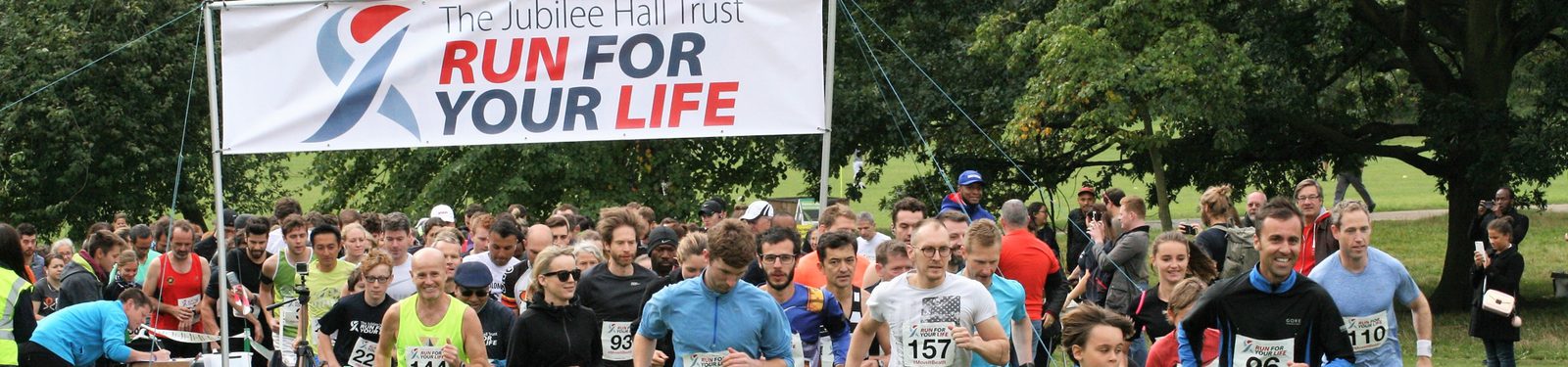 A large group of runners gathered at the starting line of a race in front of a banner that reads "The Jubilee Hall Trust RUN FOR YOUR LIFE." The background features green trees and a park setting. Participants wear various race bibs and colorful athletic attire.