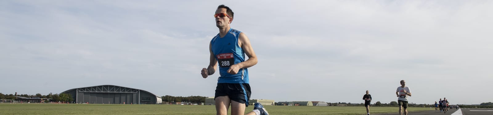 A man in a blue tank top and black shorts is running on a wide, open road during a race. He wears red sunglasses and a race bib. The background shows more runners, an airplane hangar, and an open sky.