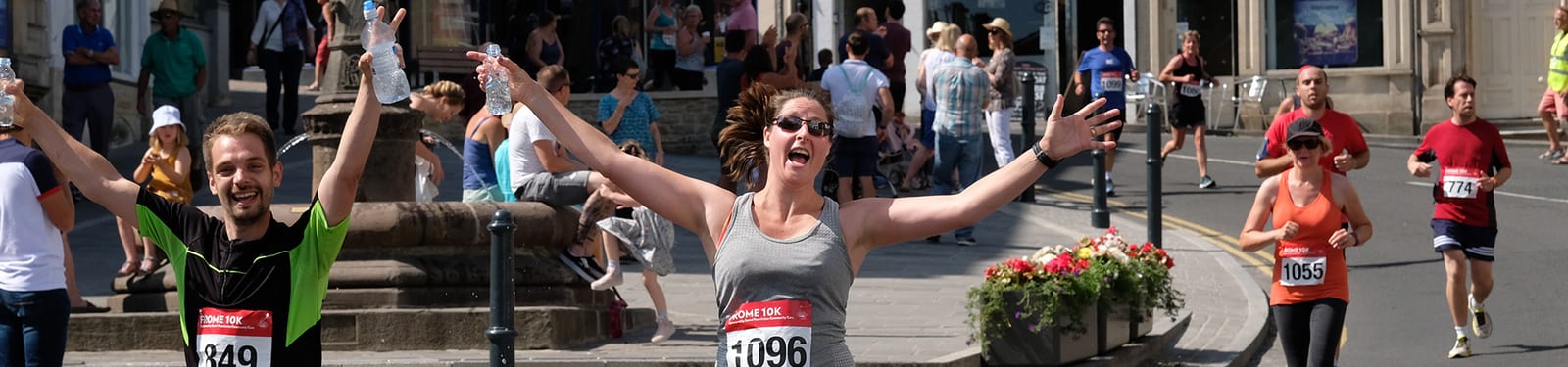 A group of runners on an urban street participate in a race. A woman with her arms raised and a big smile looks excited, alongside other runners wearing numbered bibs. Spectators sit on a ledge and stand nearby, cheering and enjoying the sunny day.