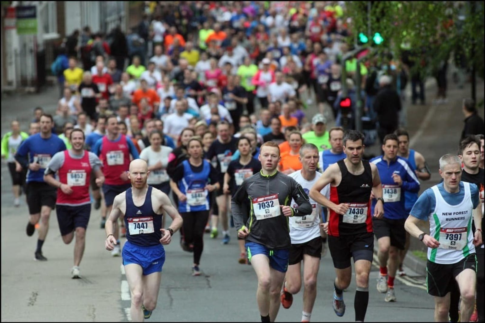 A large group of runners participating in a road race or marathon, wearing various brightly colored athletic clothing and race numbers. Some runners are in the lead while others follow closely behind. Trees and buildings line the course in the background.