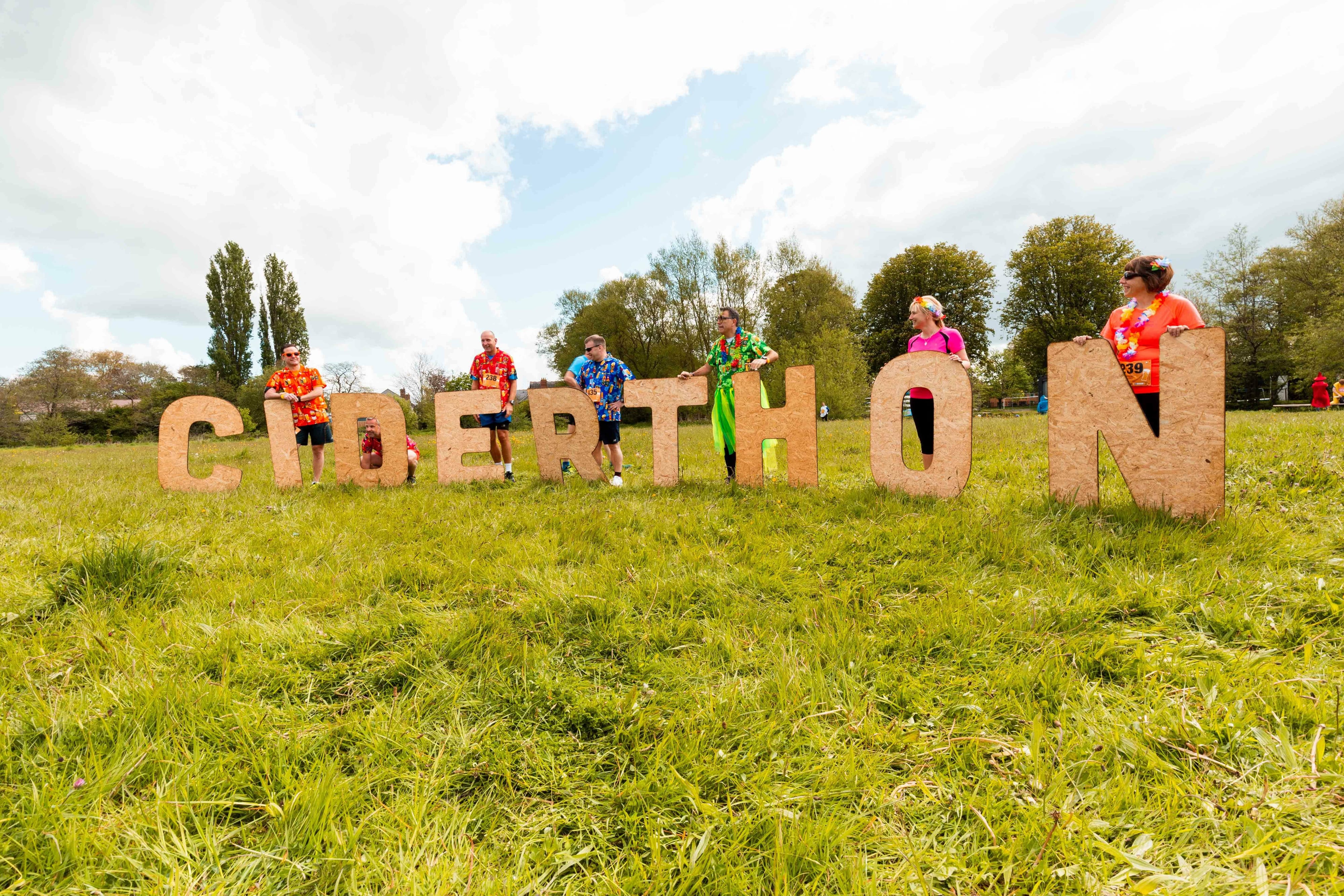 People stand behind large, upright letters spelling "CIDERTHON" on a grassy field. They are dressed in colorful outfits, and the sky is partly cloudy. Trees and other participants can be seen in the background. The atmosphere appears festive and cheerful.